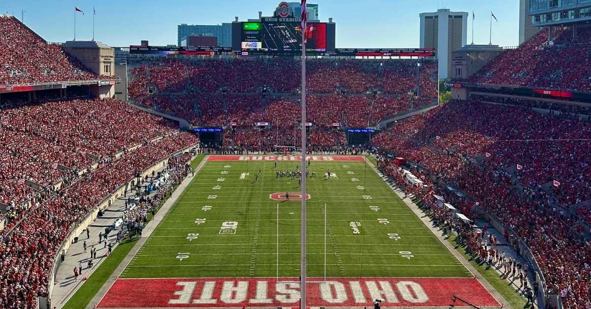 High-angle view of a crowded Ohio Stadium during an Ohio State Buckeyes football game, with a green field, red end zones labeled “OHIO STATE” and “BUCKEYES,” and fans filling the stands under a clear blue sky.