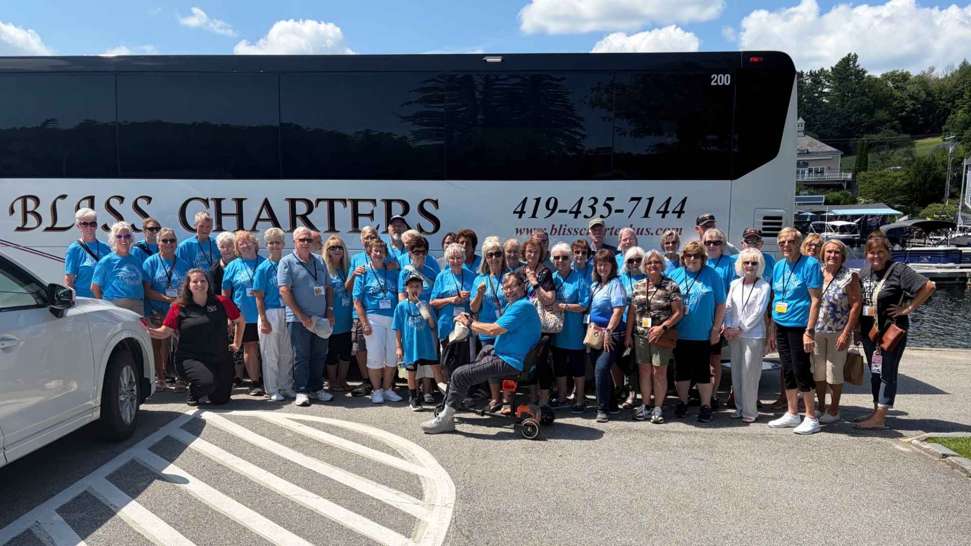 Tour Group in Front of Charter Bus: Large group of travelers, many wearing matching blue shirts, posing in front of a white Bliss Charters bus near a waterfront marina on a sunny day.